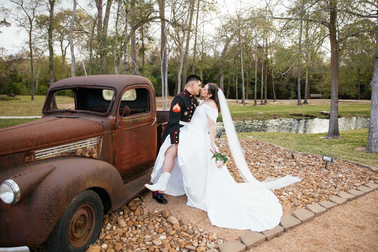 Bride with flowing veil kissing her groom beside an antique truck at a wooded Houston wedding venue
