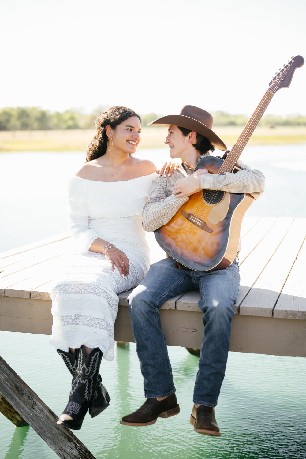 Bride and cowboy groom with a guitar sitting together on a lakeside dock in Texas